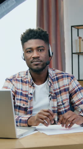 Attractive smiling successful young black-skinned guy in headphones with mic sitting in front of camera in pause between recording vlog for his audience