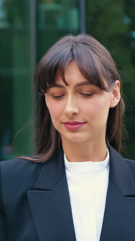 Close-up of a young woman posing for the camera and smiling, standing outdoors in front of modern commercial office building. Urban lifestyle, professional appearance, natural daylight