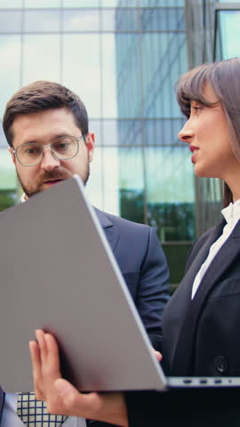 Two business professionals in formal attire standing outdoors, collaborating on remote work using laptop and tablet in a modern urban environment
