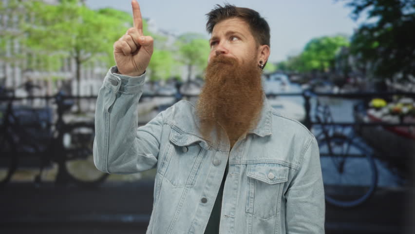 Man with long beard and denim jacket pointing finger to object on city street; inspiration enthusiasm.