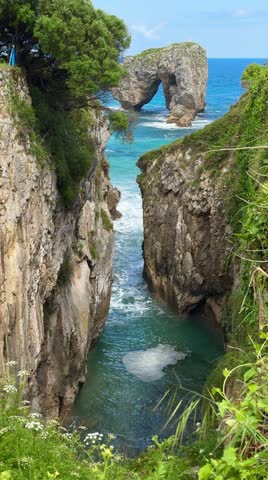 Rock arch rising from turquoise sea along rugged coastal cliffs in sunlight. Llanes, Spain