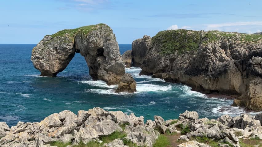 Rock arch rising from turquoise sea along rugged coastal cliffs in sunlight. Llanes, Spain