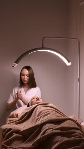 Professional cosmetologist performing a skincare facial treatment in a spa or beauty salon. A young woman is lying under a blanket while receiving relaxing facial care under special salon lighting