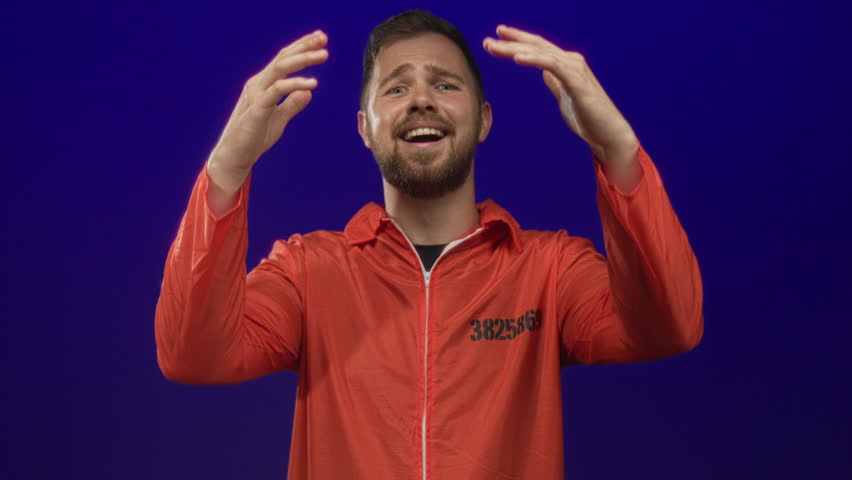 Bearded young prisoner man in orange jumpsuit with hands on head in blue studio; surprise redemption.