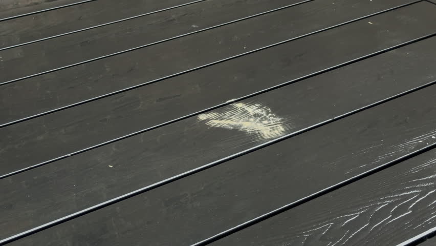 Closeup of water being thrown over some stain mess on the flat surface of outside composite decking boards, followed by a broom sweeping the excess water away.