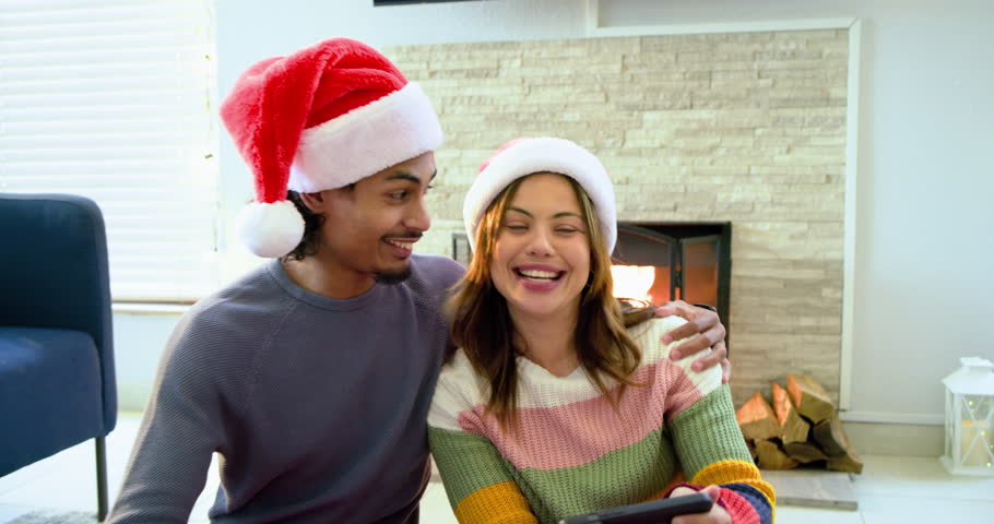 Man and woman wearing Santa hats raising smartphone for selfie laughing at surprises by fireplace. Festive, cozy, candid, warmth, companionship, celebration, lifestyle
