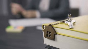 Dolly shot of property keys with a wooden house keychain on notebooks, with a female real estate agent working in the background, illustrating home ownership. - Powered by Shutterstock - Get 15% off with code: PIKWIZARD15
