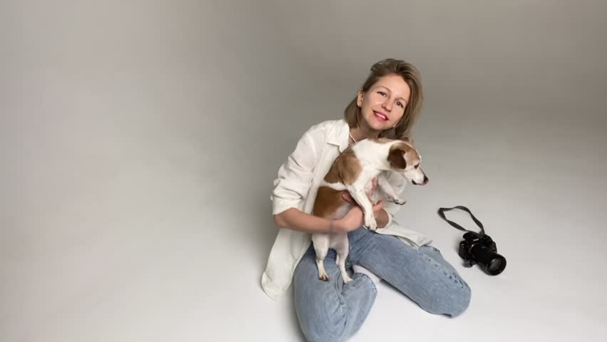 blonde woman in jeans and white shirt sits on floor hugging small happy dog Jack Russell terrier ​​and laughing. Happy moments with pet. Dog kissing and wags tail happily. Professional photo camera 