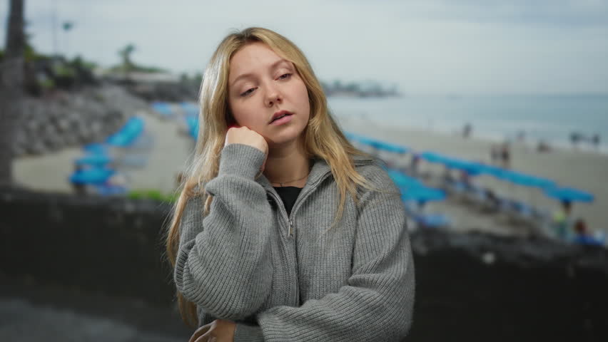 Woman with blonde hair in thoughtful pose at the seaside, standing near a beach with blue umbrellas under a cloudy sky, dressed in a casual gray sweater.