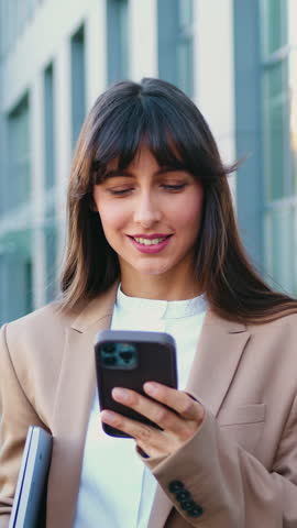Portrait of a smiling young business woman using smartphone and looking at the camera while standing outdoors in front of a modern office building