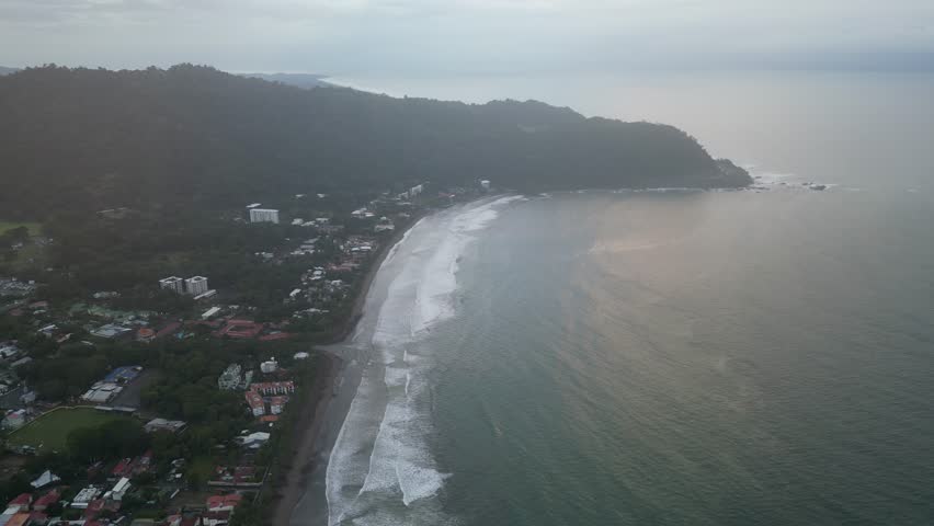 Vertical aerial shot of a coastline full of tourists with waves at dawn, with tourist and hotel buildings and businesses on Jacó Beach located in the province of Puntarenas in Costa Rica