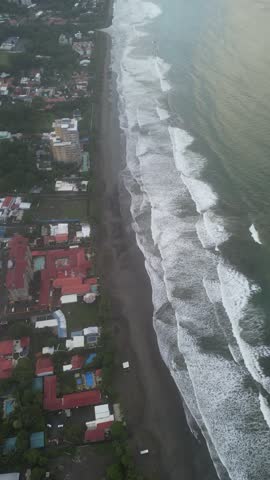 Vertical aerial view of the coastline filled with tourists, with waves in the mountains, buildings, and tourist and hotel businesses on Jacó Beach located in the province of Puntarenas in Costa Rica