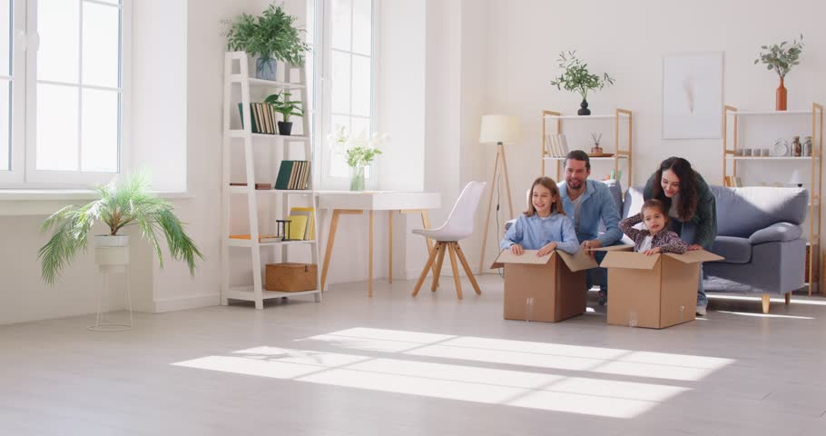 Parents engage children in playful activity by rolling them around living room in cardboard boxes. Brother and sister sit in boxes while mother and father playfully push them across living room floor.