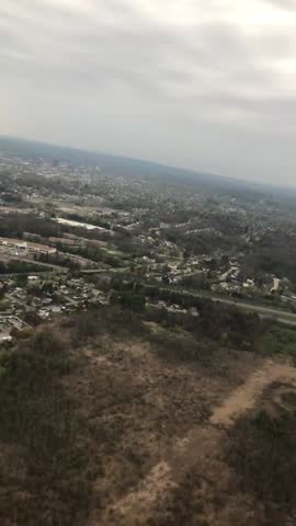 Aerial ascent over suburban Manchester New Hampshire with dense mixed forest, retail corridors, and early-spring leaf-off landscape under overcast stratiform cloud deck