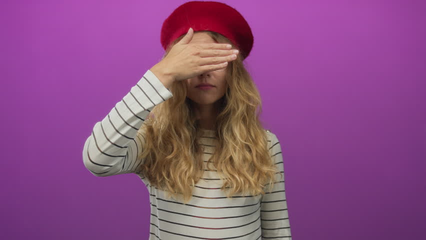 Woman places hand covering eyes in a studio portrait of a blonde young model wearing a red beret and glasses against a purple background.