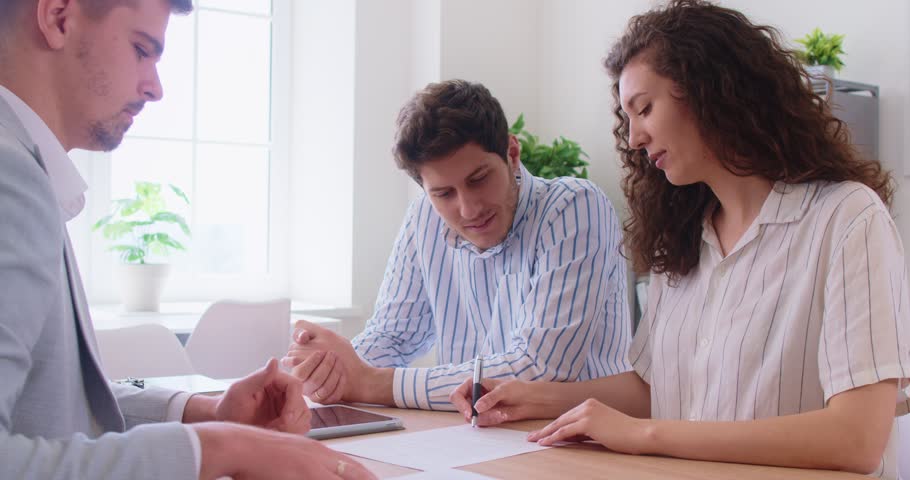 Positive couple signing contract with realtor in office for new home purchase, finalizing real estate deal, engaging with agent, radiating enthusiasm, completing property transaction with confidence.