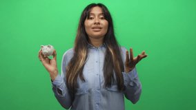 Woman brunette holding small white piggy bank in green studio while closing eyes and making a disappointed face; financial frustration. - Powered by Shutterstock - Get 15% off with code: PIKWIZARD15