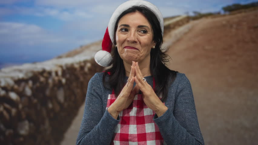 Woman in colorful red santa hat and checkered apron shaping heart with hands in studio; festive affection.