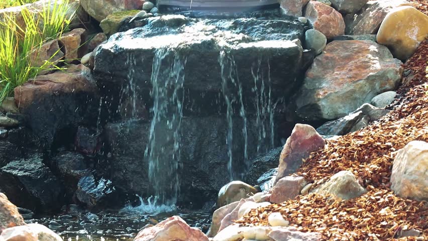 A close up view of a small man made waterfall in a garden. Water flows over dark rocks into a stream surrounded by mulch and plants, creating a serene and relaxing scene.