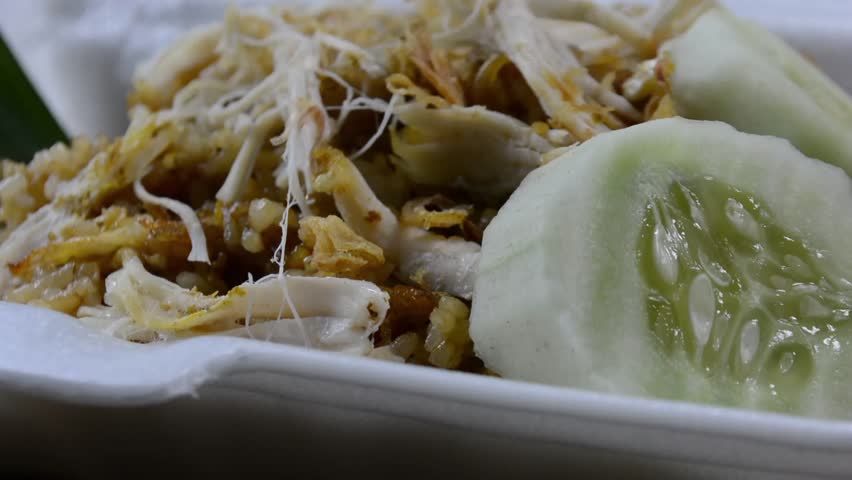 Close-up of Nasi Goreng Kampung (Indonesian fried rice) topped with shredded chicken (Ayam Suwir) and cucumber slices on a foam container