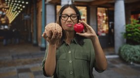 Young woman with glasses and brunette hair holds a model brain in one hand and a red heart in the street, frowning at both; mind versus heart conflict. - Powered by Shutterstock - Get 15% off with code: PIKWIZARD15