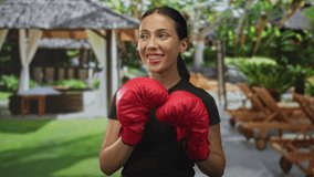 Young hispanic woman boxer wearing red boxing gloves, fists raised in front of a building poolside gazebo, smiling with eyes closed; joy fitness. - Powered by Shutterstock - Get 15% off with code: PIKWIZARD15