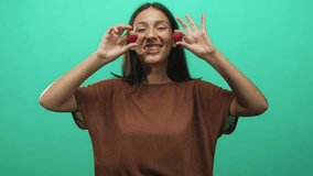 Young brunette woman smiles and holds strawberries beside cheeks in studio with green backdrop; playful joy. - Powered by Shutterstock - Get 15% off with code: PIKWIZARD15