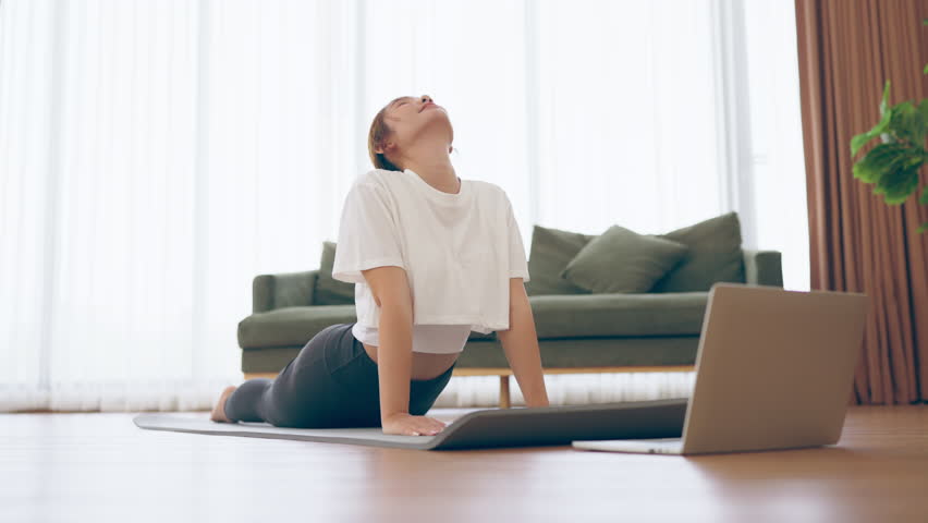 Young Asian woman stretching yoga workout on exercise mat while online training class with computer laptop in living room