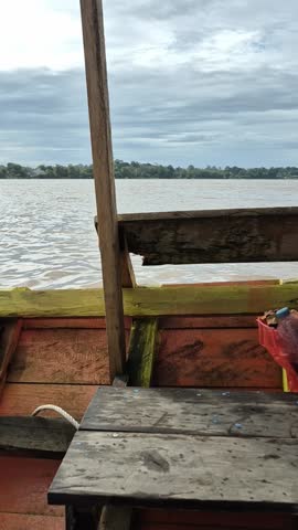 A serene river view from a wooden boat, with calm waters stretching to the horizon under a cloudy sky.