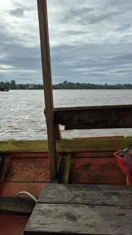 A serene river view from a wooden boat, with calm waters stretching to the horizon under a cloudy sky.
