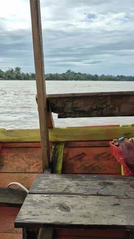 A serene river view from a wooden boat, with calm waters stretching to the horizon under a cloudy sky.