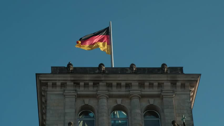 Dynamic footage of the German national flag waving against a clear blue sky above the historic Reichstag building, the seat of the Bundestag in Berlin. Captures the symbolism of democracy, government,