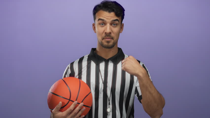 Hispanic man dressed as a basketball referee holding a ball against an isolated purple background, pointing to himself confidently.