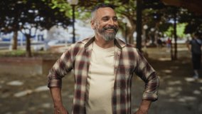 Man with grey hair and beard smiling with hands on hips wearing a plaid shirt on a tree lined street near park benches; contentment. - Powered by Shutterstock - Get 15% off with code: PIKWIZARD15