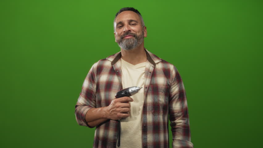Man carpenter holding power drill, right hand visible on tool, smiling, middle aged with grey hair and beard in green studio; friendly confidence.