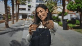 Woman points finger at viewer while holding takeaway coffee cup wearing denim apron on street; welcoming local cafe. - Powered by Shutterstock - Get 15% off with code: PIKWIZARD15