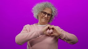 Woman with curly grey hair in glasses and pink sweater stands against isolated pink background making a heart shape with her hands, exuding warmth and positivity. - Powered by Shutterstock - Get 15% off with code: PIKWIZARD15