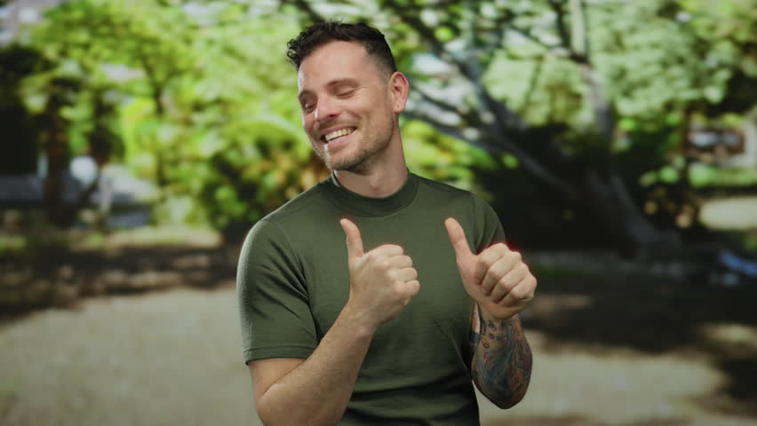 Young man smiling with thumbs up in a sunlit park showcasing a cheerful and confident expression outdoors during a bright day surrounded by lush green trees.