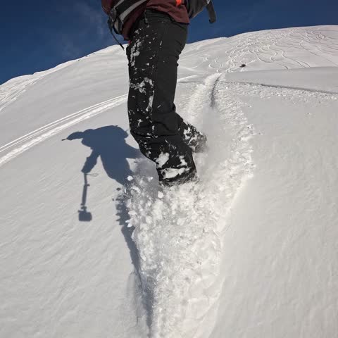 Extreme close-up POV from the front, showing the snowboarder's lower body and gear creating dynamic snow spray as they execute a fast, aggressive turn on the slope
