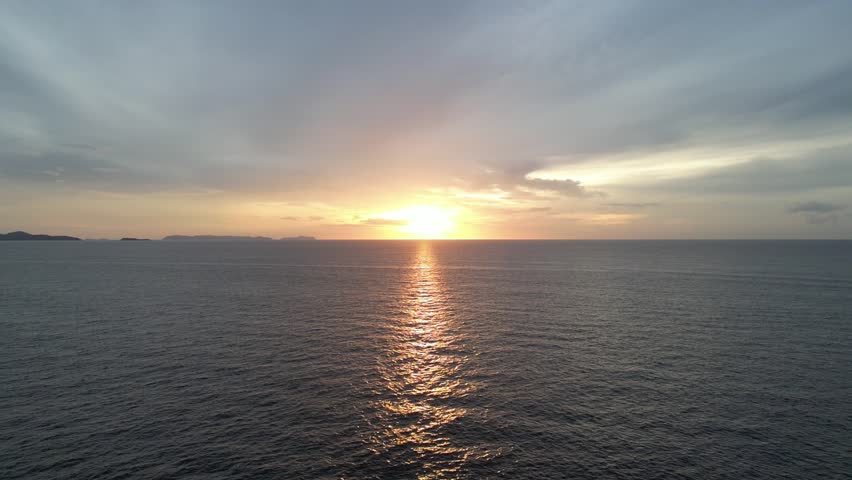 A drone view of a dramatic sunset over a dark ocean, with the sun's reflection creating a shimmering path on the water.