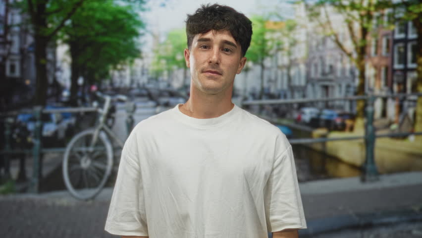 Man, young hispanic, makes heart gesture with hands on street in amsterdam wearing white tshirt and looking at camera; love connection.