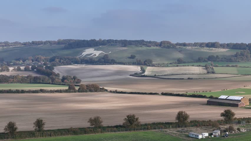 A wide aerial view of the English countryside, showcasing rolling farmland, green pastures, and the iconic white chalk horse carved into the hillside