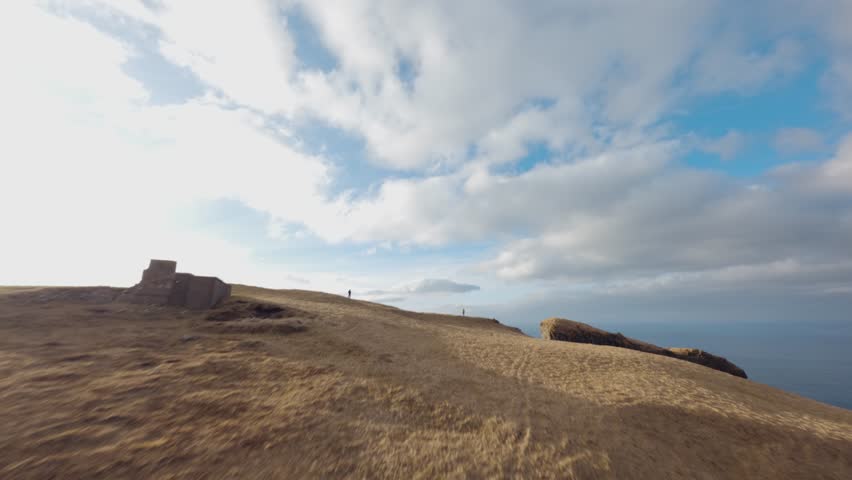 A drone view of person in yellow jacket standing near cliff edge by Eioi, Faroe Islands, above rugged coast and bright sky