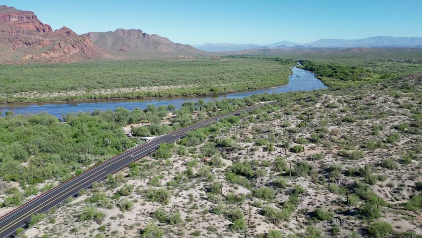 An aerial view of the Salt River cutting through Arizona's desert valley, with red cliffs, green vegetation, and open highway beneath a clear sky
