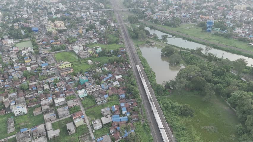 A smooth 4K 10-bit 60fps drone city-revealing shot showing a wide suburban town, railway tracks, greenery, scattered houses, and a calm lake captured in soft evening light.
