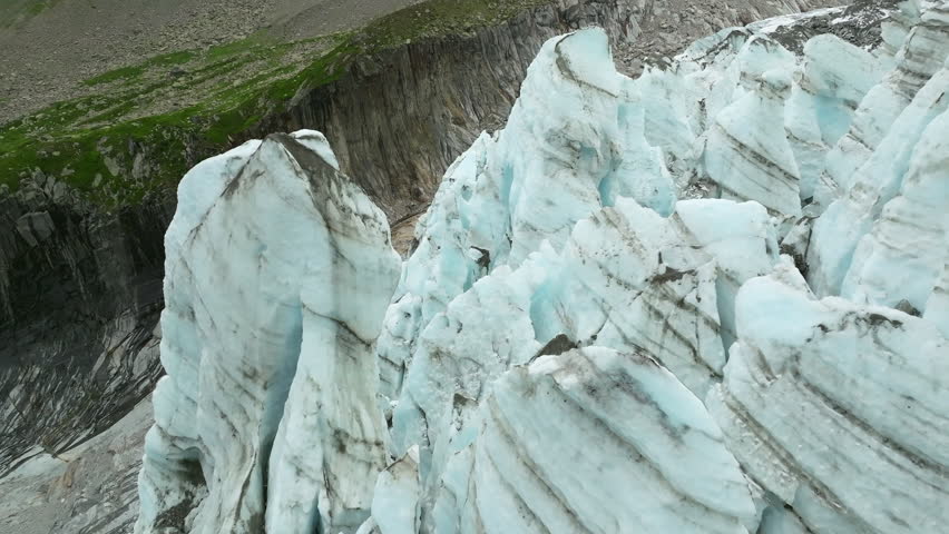 A closeup, drone shot of towering blue ice pinnacles on the Argentiere Glacier contrasting with steep rocky cliffs in the French Alps