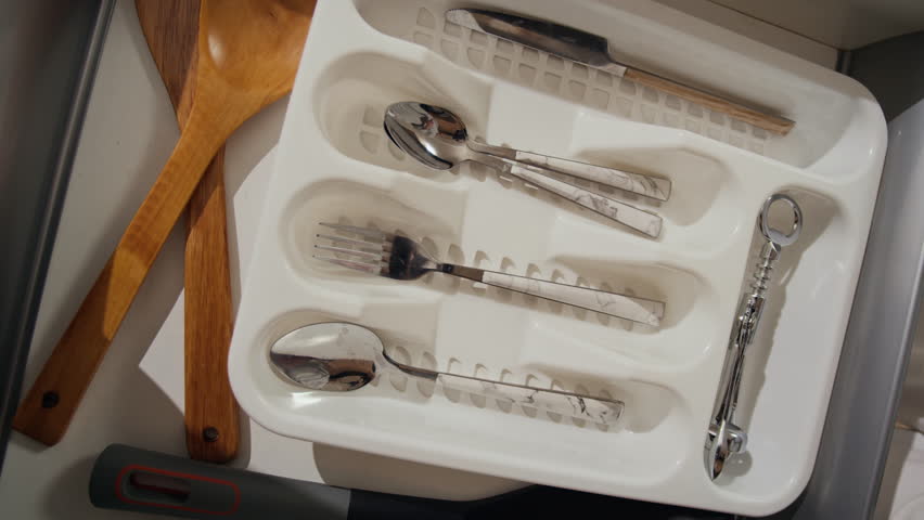 High angle shot of clean and polished cutlery including spoons, forks, table spoon in drawer organizer. Unrecognizable female hand putting just washed spoon in it