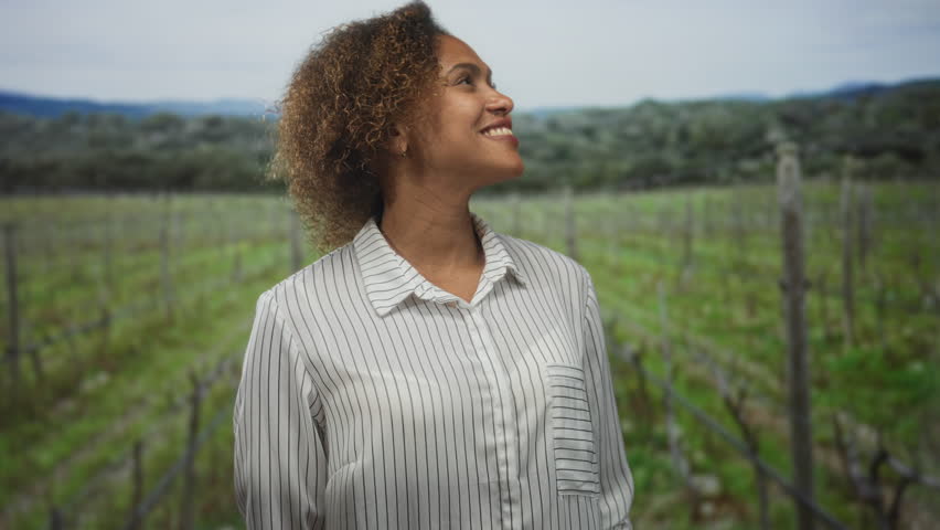 Woman smiling and looking up in a vineyard field, wearing a striped shirt with hands behind back and relaxed posture; joy openness calm.