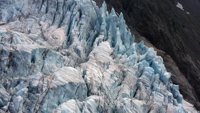 An aerial view of Trift Glacier's surface, showing intensely cracked, blue and white ice with curved flow lines next to dark rough rock in Switzerland