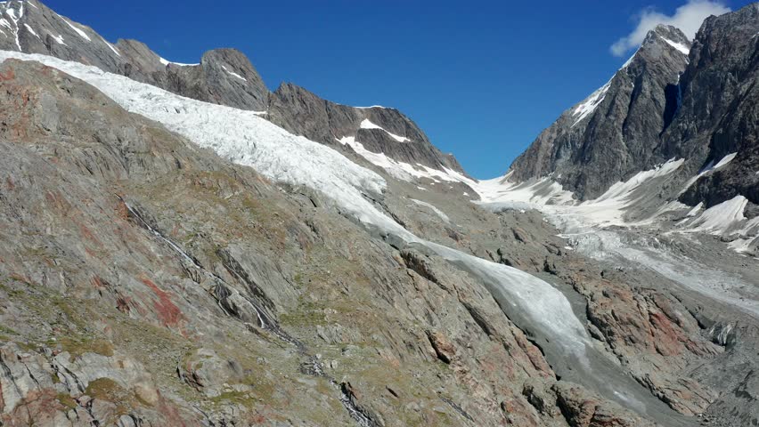 An aerial view of a Swiss glacier, showing the sloping ice flow carving through rugged cliffs and rocky terrain under bright sunlight and blue skies